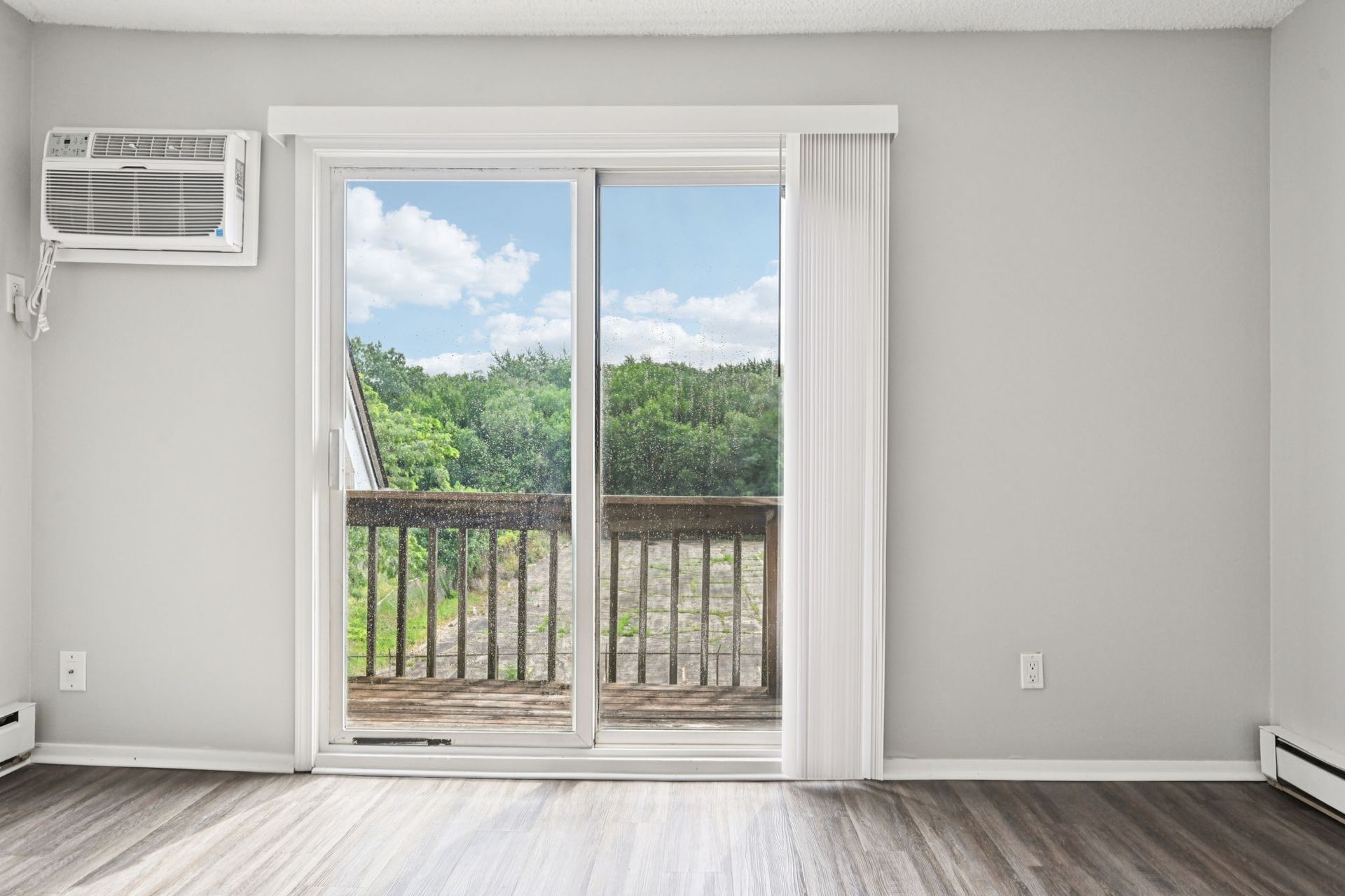 Sliding glass door opens to a wooden deck with trees. AC unit on the wall, light gray walls, dark wood floor.