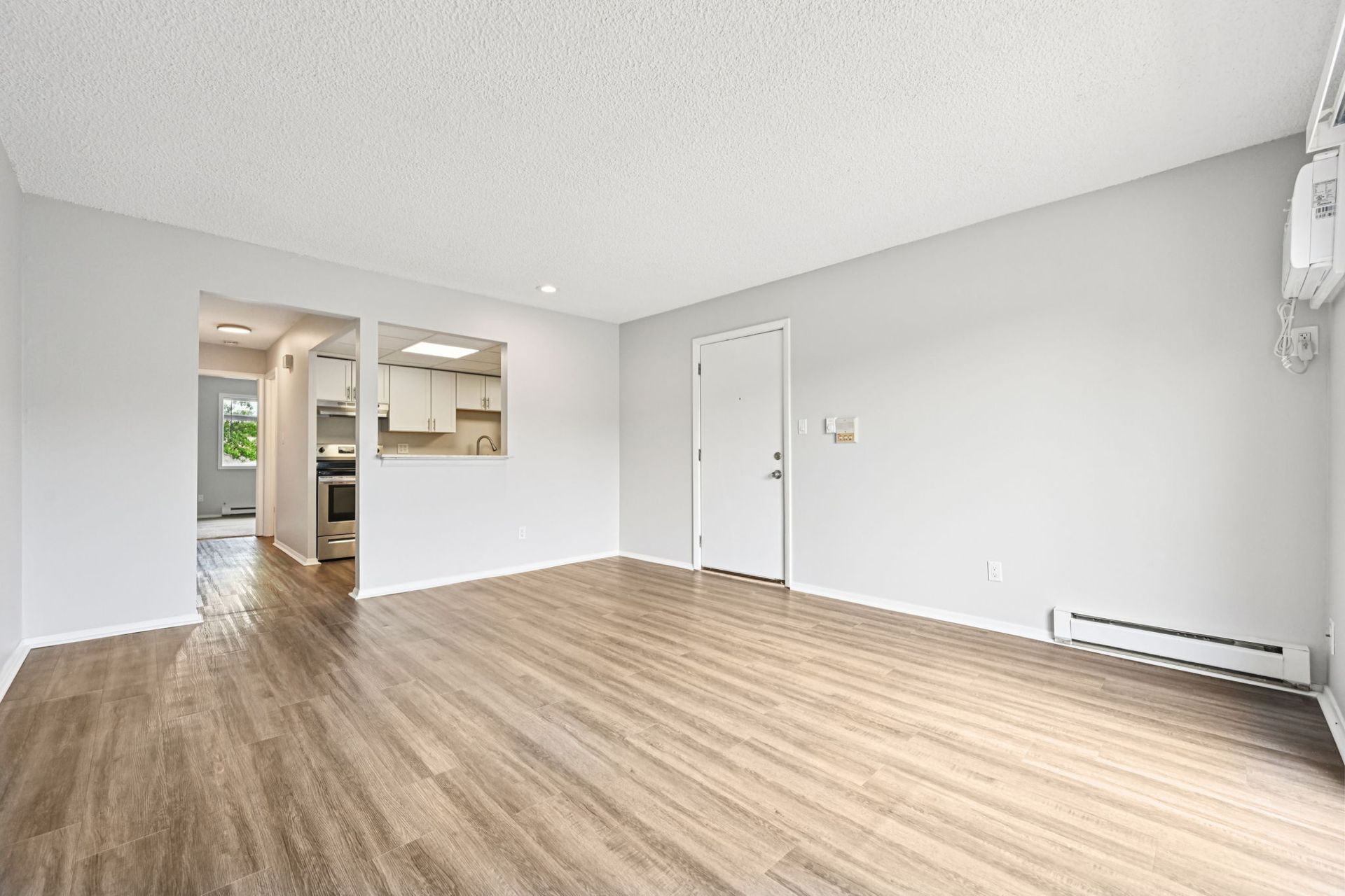 Empty living room with light wood floor, pale grey walls, and doorway to a kitchen.