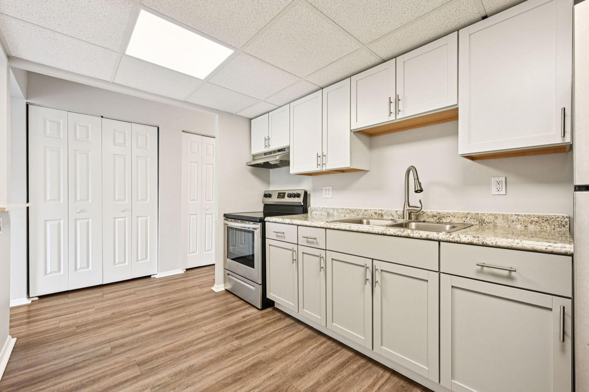 White kitchen with light gray cabinets, stove, sink, and closed pantry doors. Brown wood-look flooring.