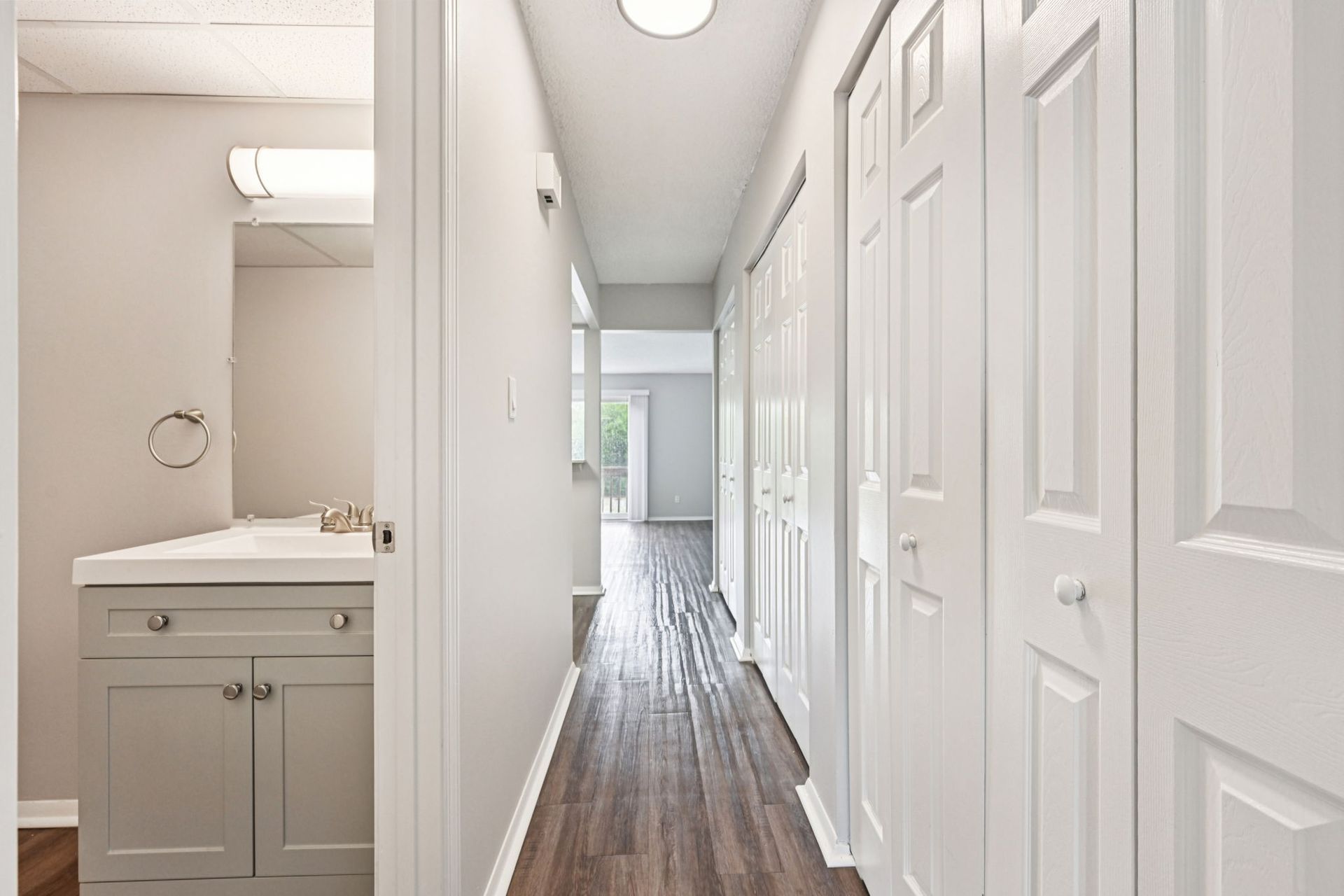 Narrow hallway with white doors, light gray walls, and dark wood-look flooring, leading to an open doorway.
