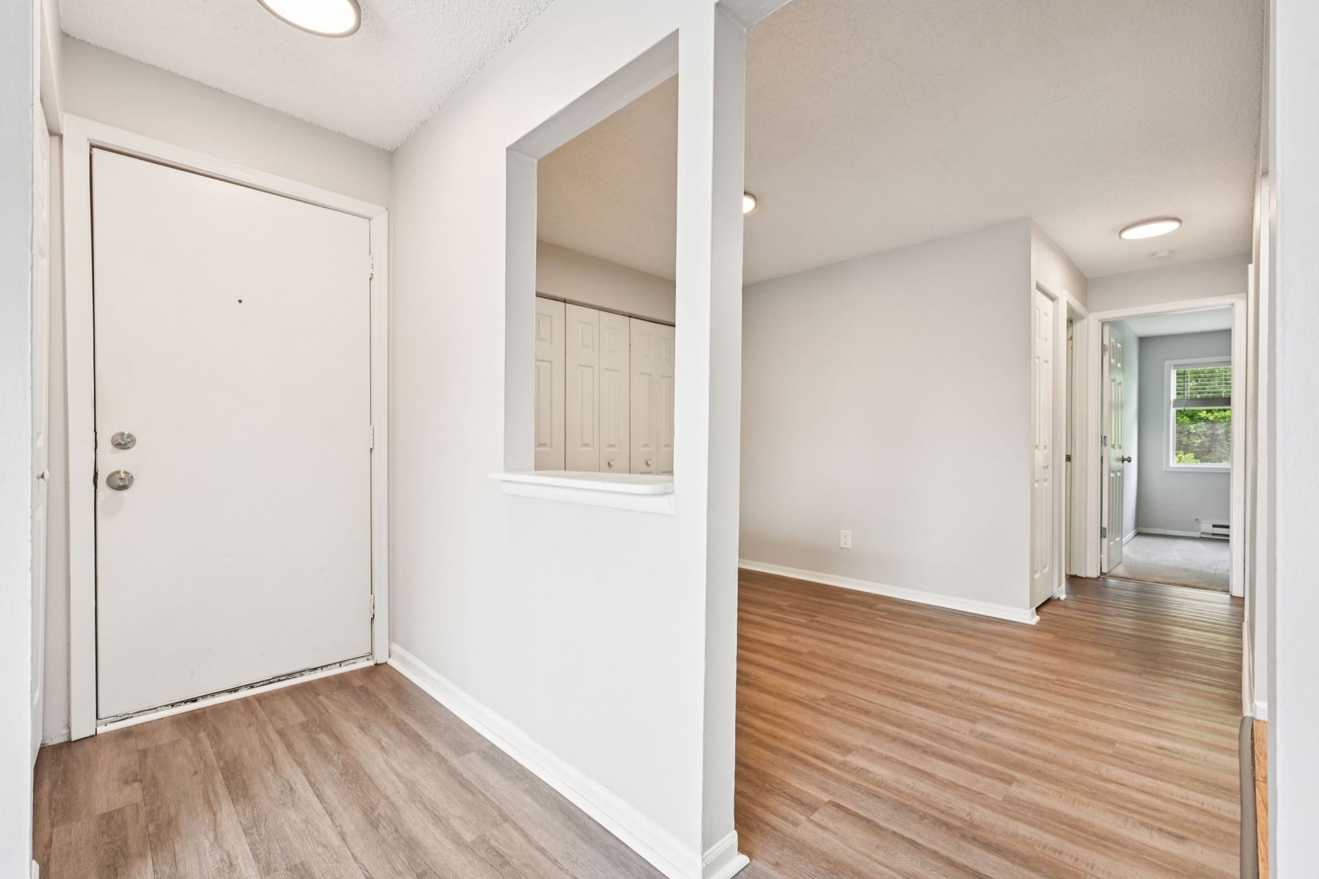 Bright apartment entryway with light wood flooring, white walls, and a hallway to the right.