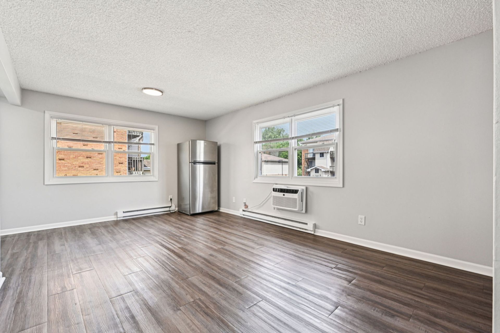 Empty apartment interior with wood floors, stainless steel refrigerator, and two windows.