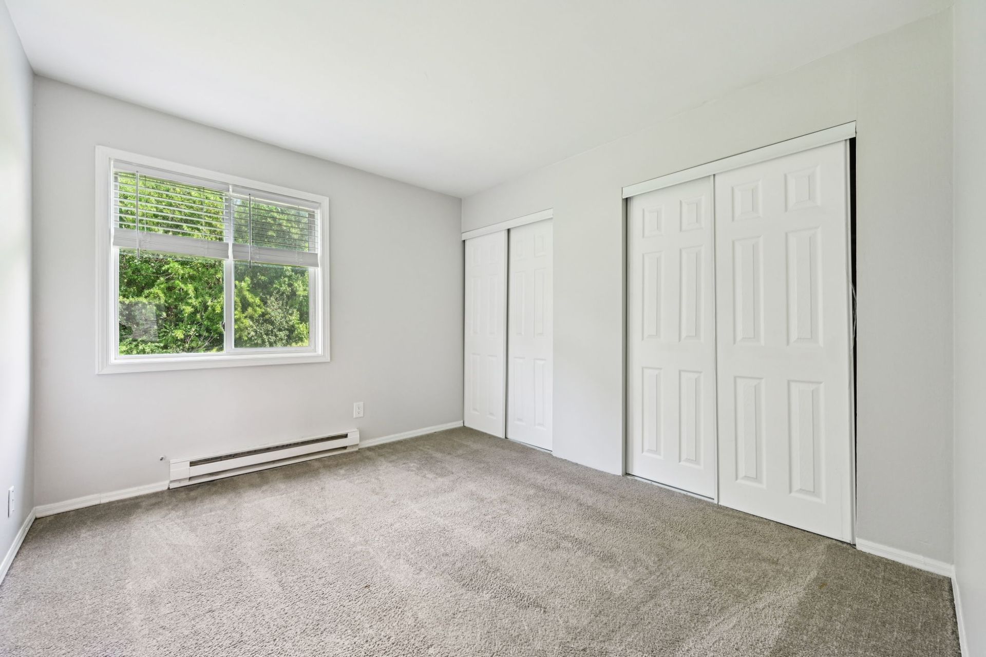 Empty bedroom with light grey walls, carpet, window, and white sliding closet doors.