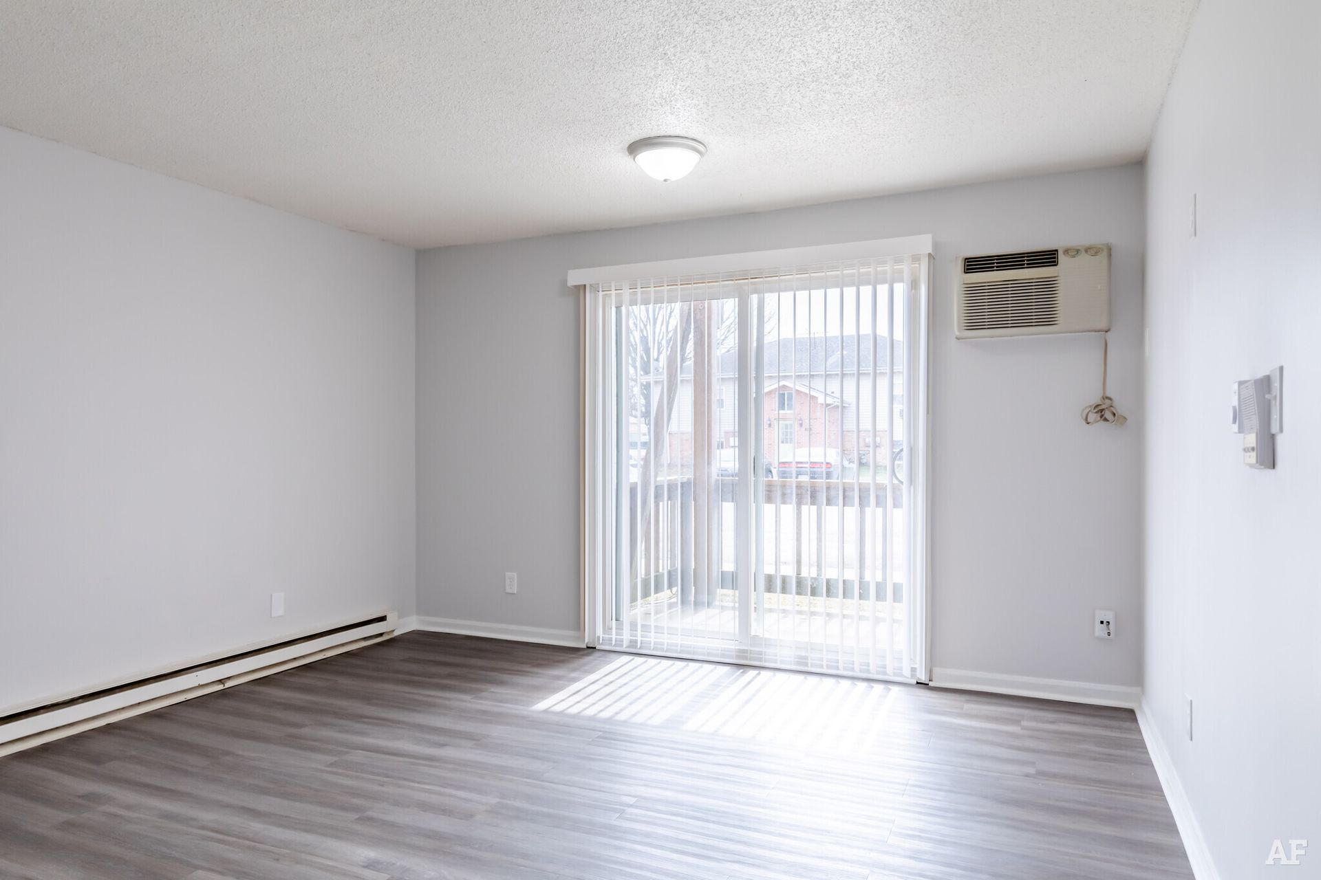 Empty living room with sliding glass door, gray walls, and wood-look floor. An air conditioner is mounted by the door.