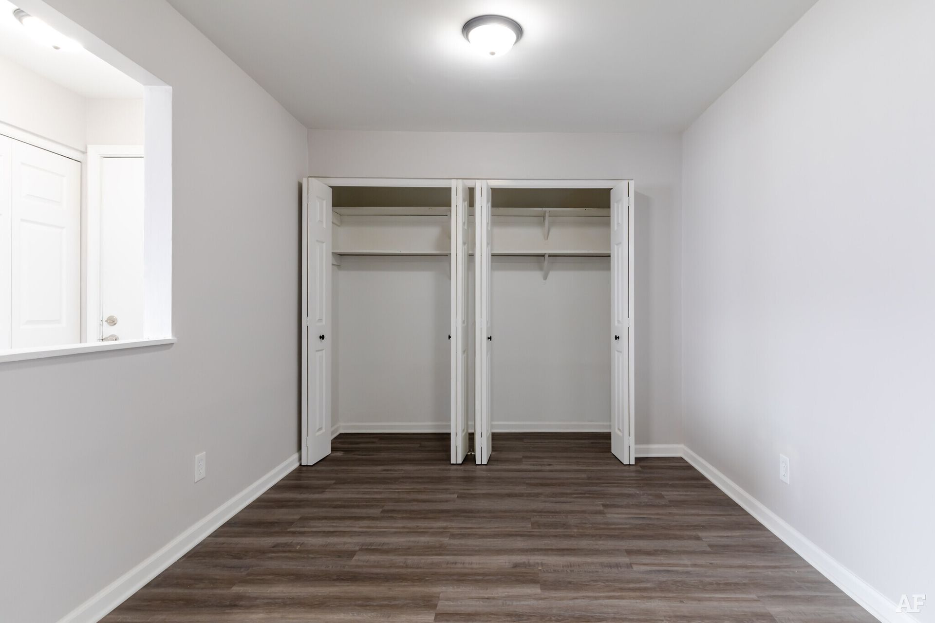 Empty room with white walls, dark wood floor, open closets, and a window.