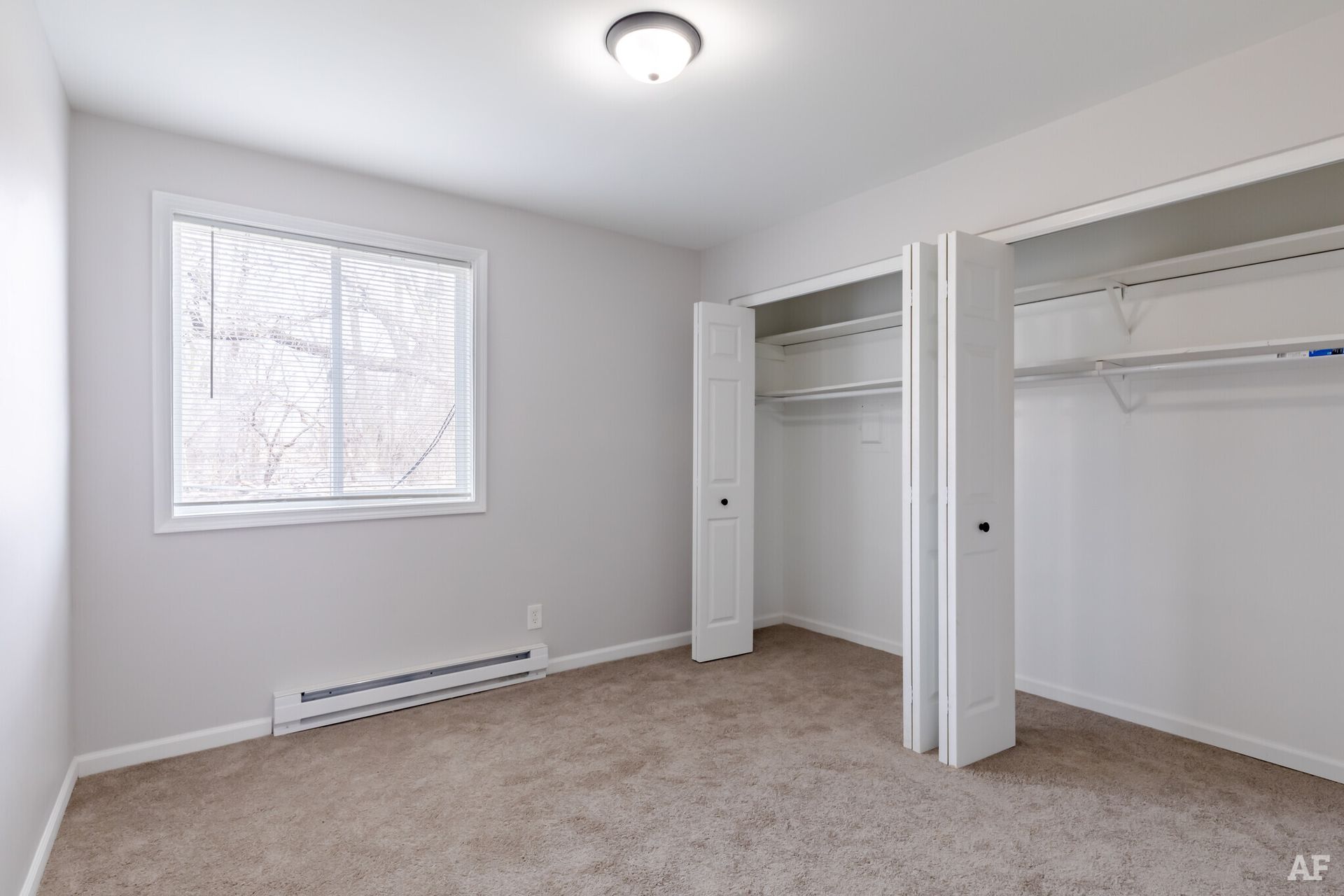 Empty bedroom with a window, closet, and tan carpet. White walls and doors.