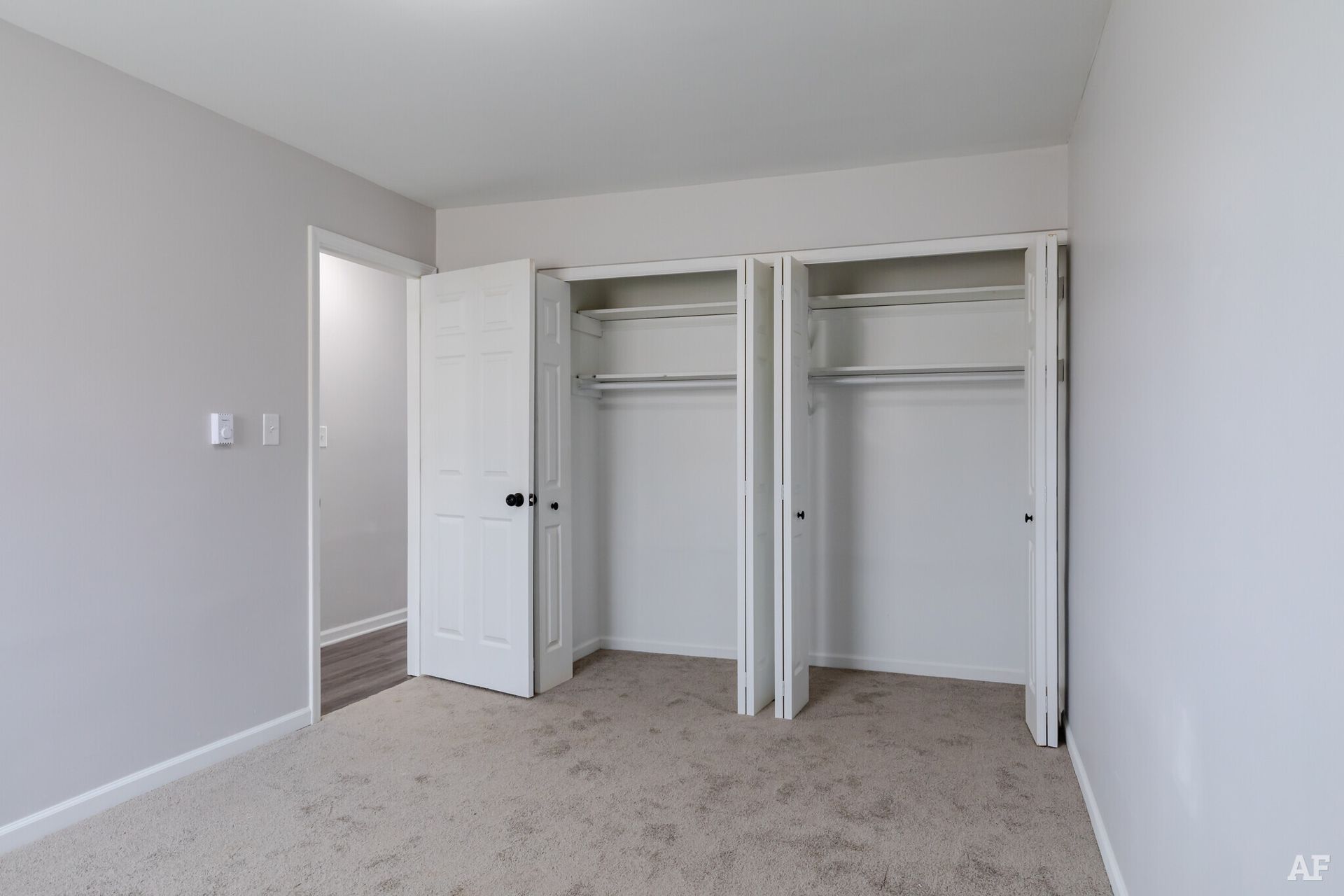Empty bedroom with gray walls, carpet, and a double-door closet. A doorway leads to another room.
