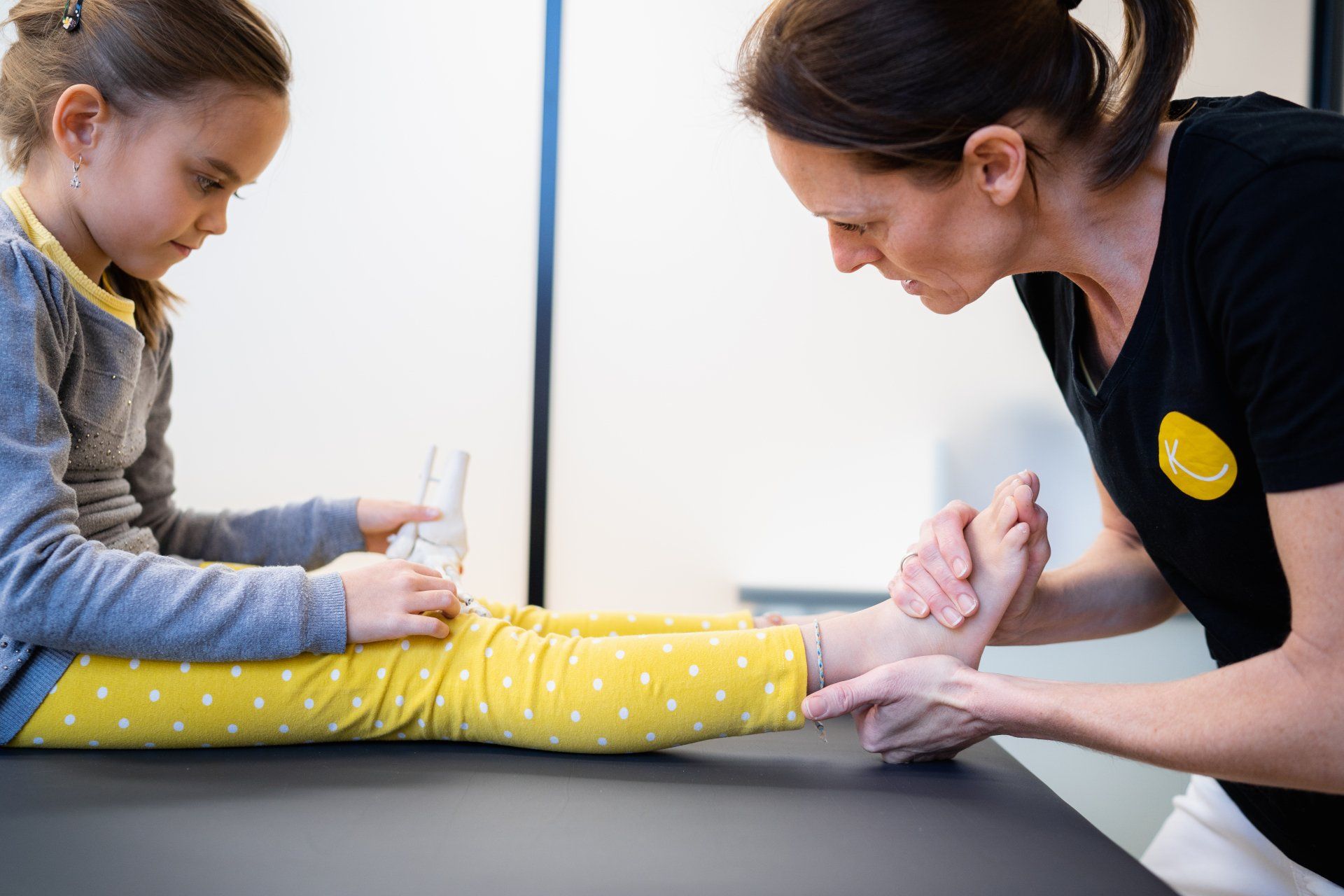 Nora Giger, MSPhT mit einem gelben Smiley auf ihrem Hemd untersucht den Fuß eines kleinen Mädchens