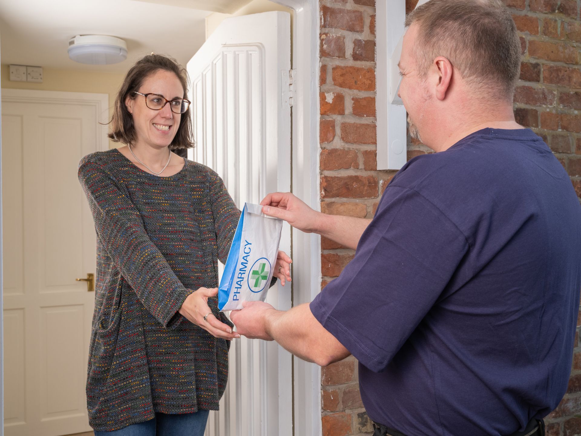 Pharmacy delivery service by male employee, handing prescription to a woman at her home.