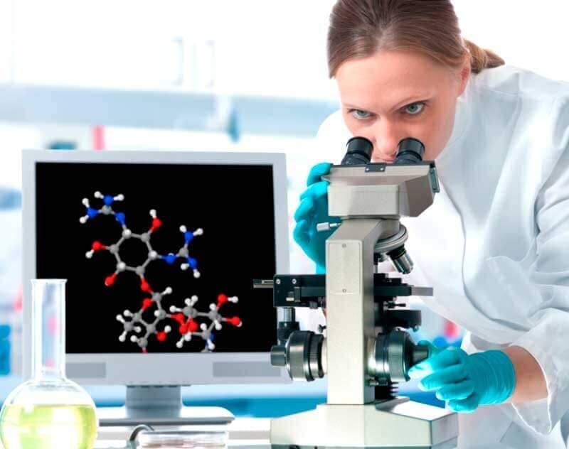 Una mujer está mirando a través de un microscopio frente a un monitor de computadora.