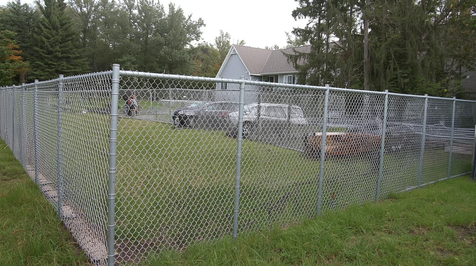 Chain-link fence encloses a yard with cars, trees, and a house in the background.