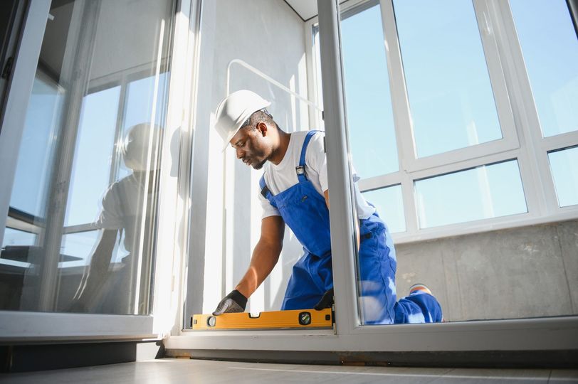 Construction worker in blue overalls and hard hat using level on window frame.