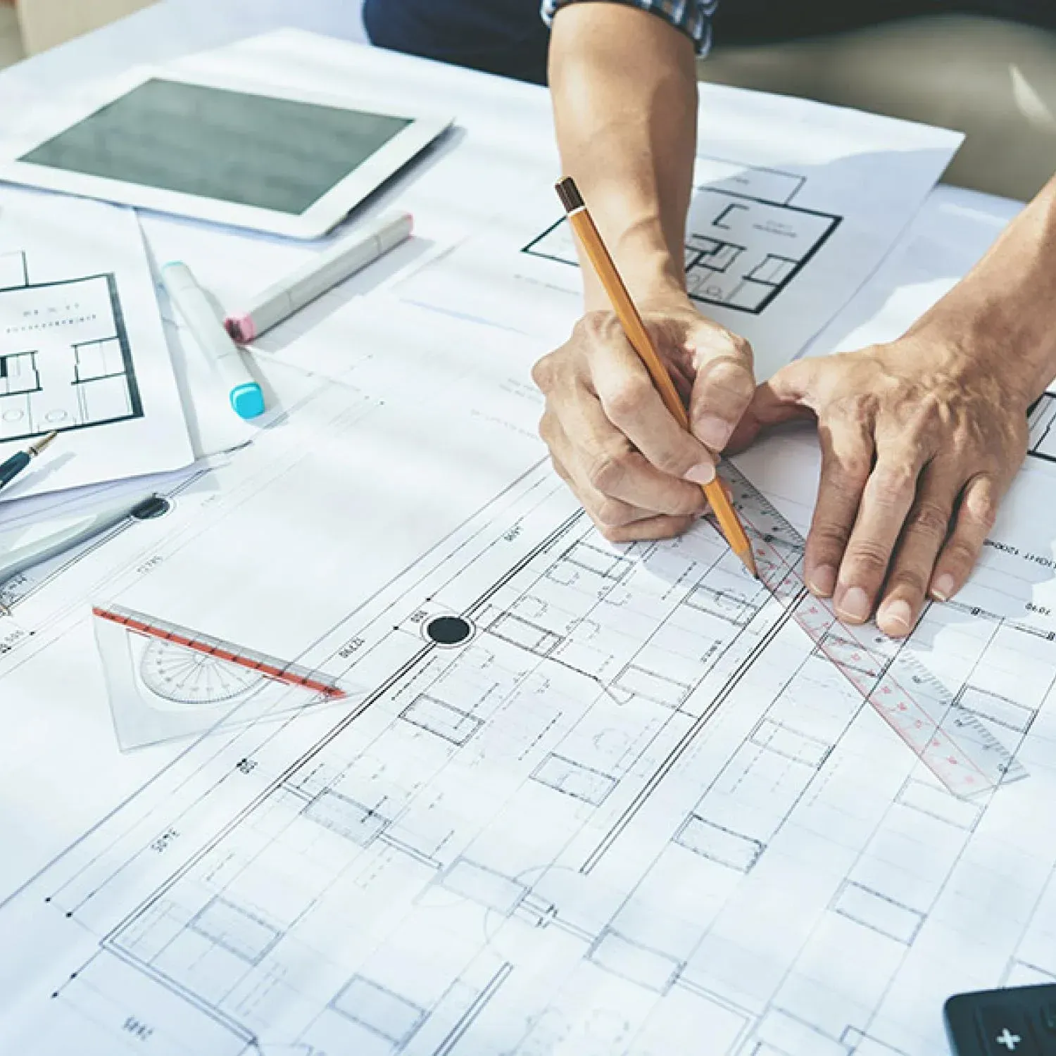 Person using a pencil and ruler to draw on architectural blueprints at a desk with a tablet and other tools.
