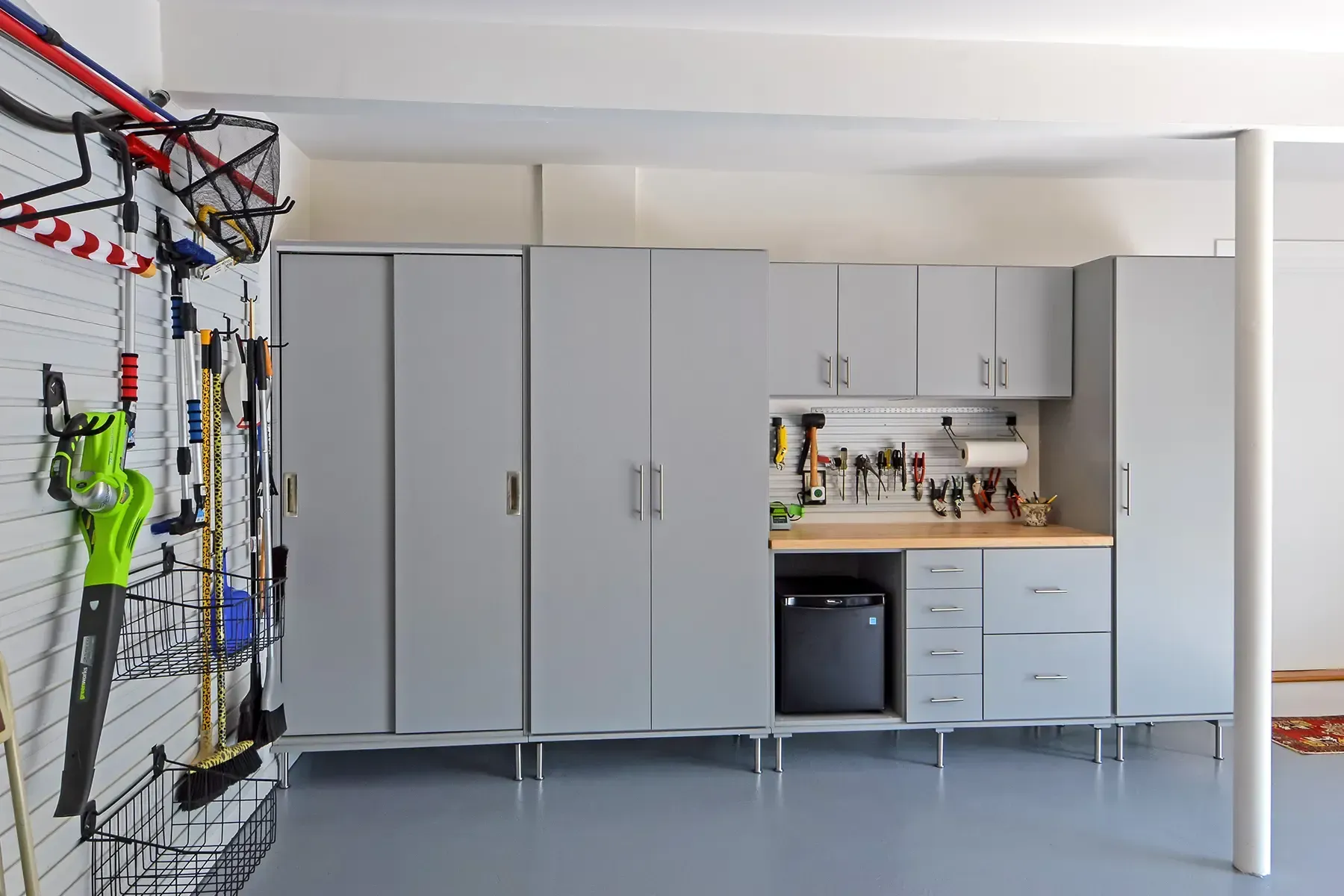 Gray garage with organized storage cabinets and tools. Pegboard wall, work surface, and mini-fridge.