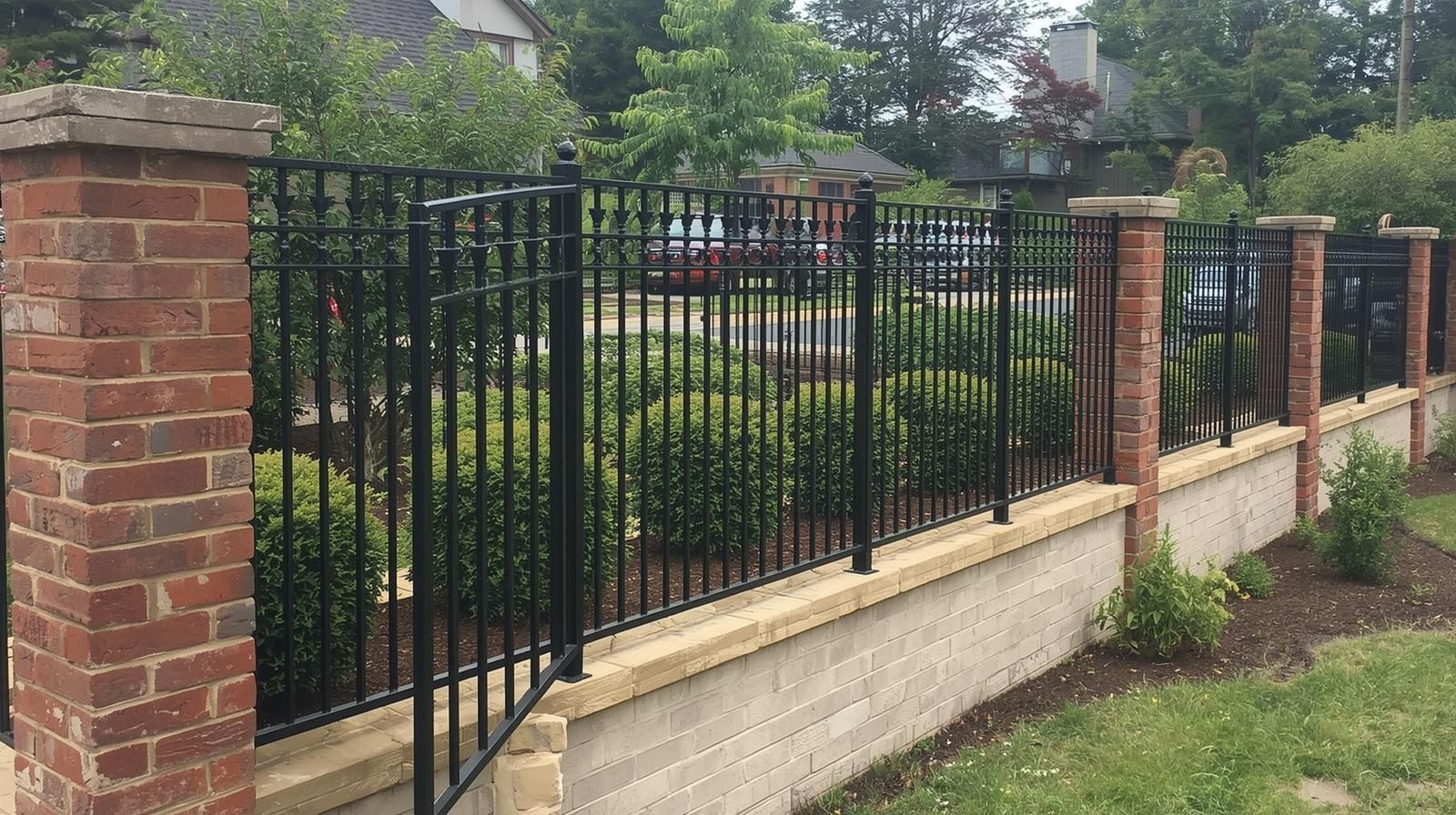 Brick and black metal fence with green shrubbery, on a lawn with plants and trees.