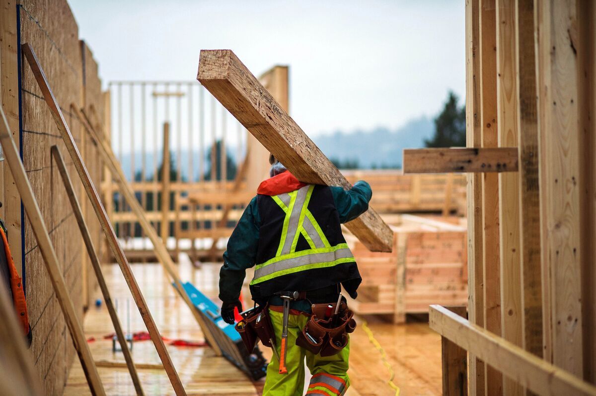 Construction worker carrying wood beam on shoulder, wearing high-visibility vest. Building frame in progress outdoors.