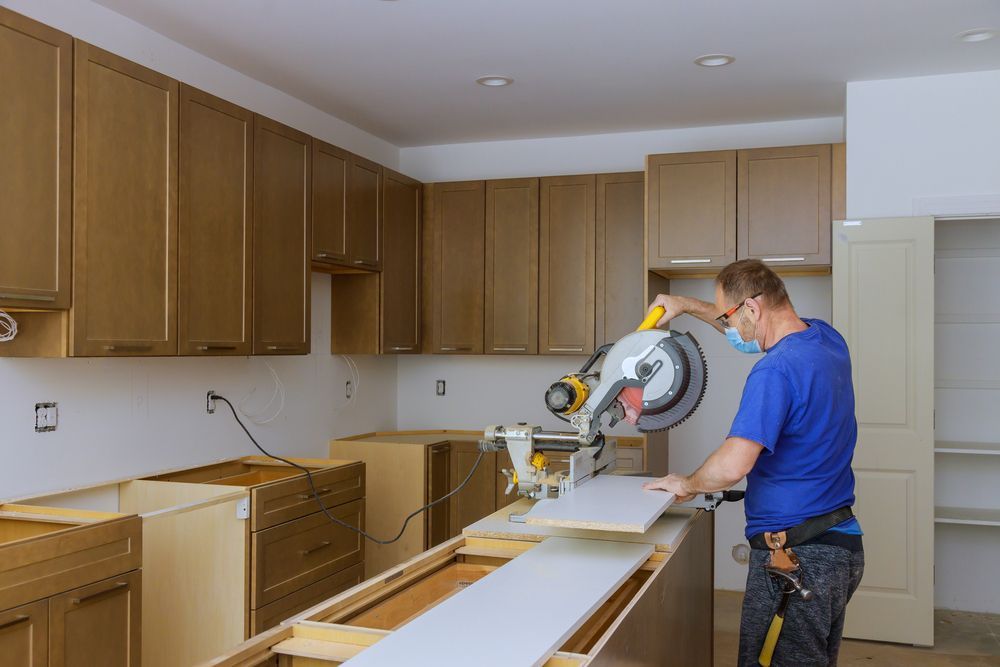 Man in blue shirt and mask cutting wood with a saw in a kitchen with new cabinets.