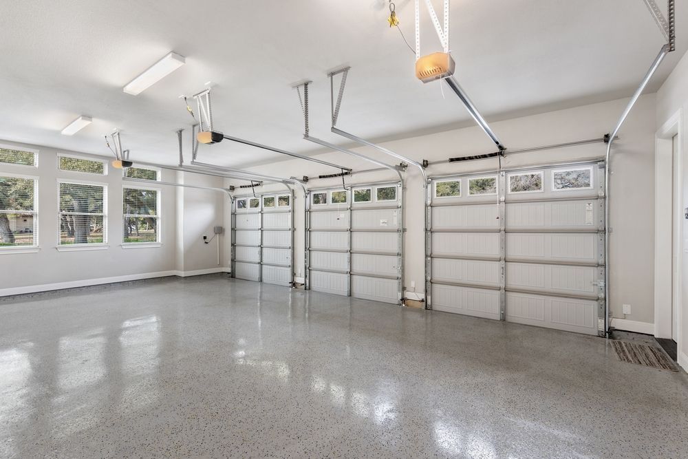 Empty three-car garage with gray epoxy floor, white doors, and large windows.