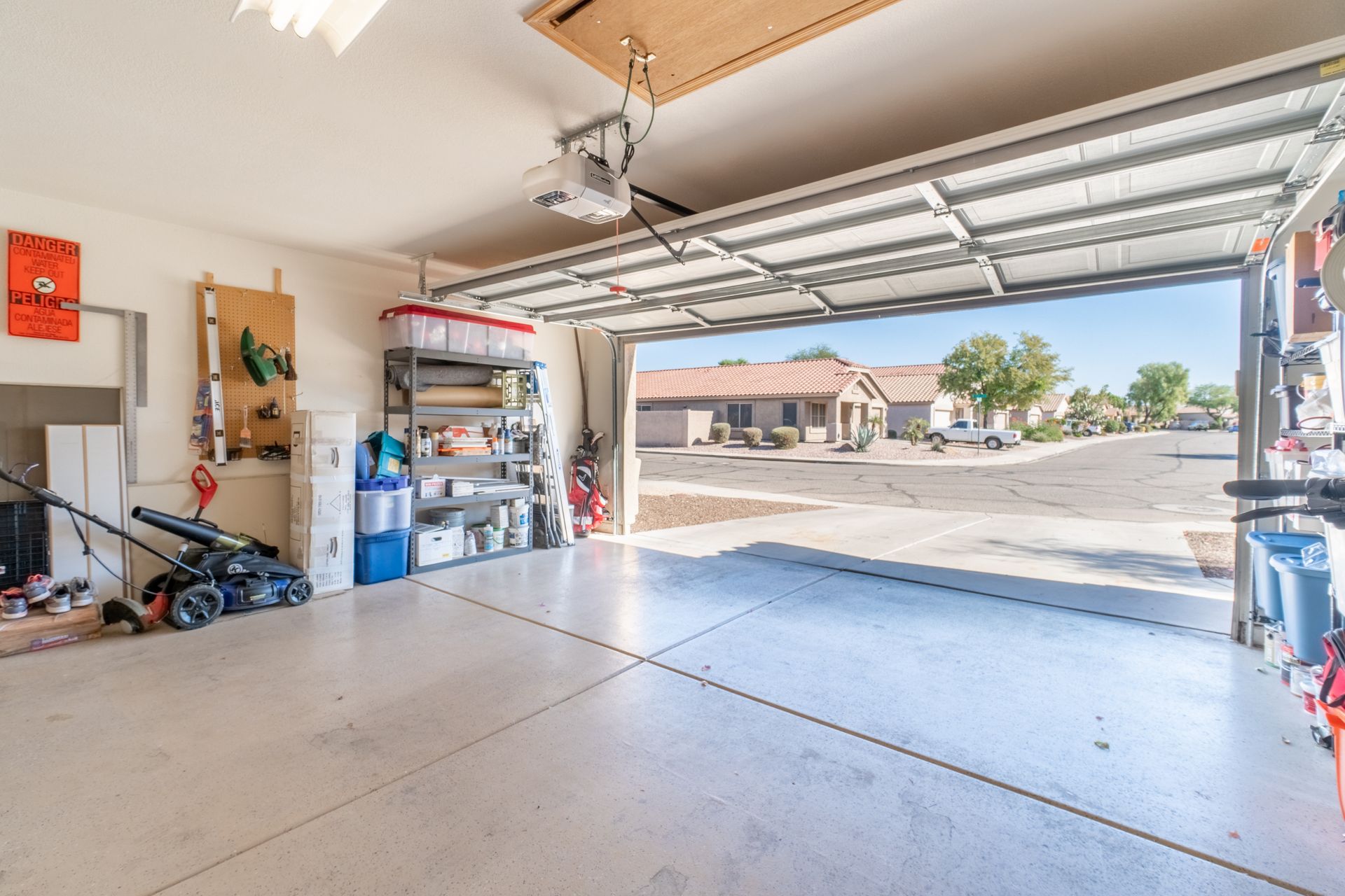 Open garage with driveway view; tools and storage on shelves, lawnmower to the left.
