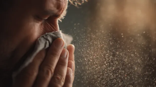 Man sneezing into a cloth, dust particles visible in the air.