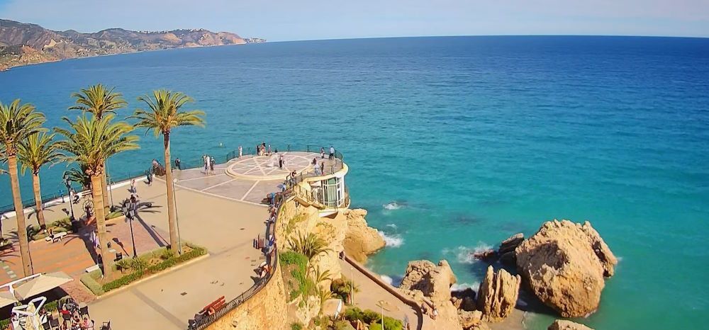 An aerial view of a beach in Nerja with palm trees and a large rock in the middle of the ocean.
