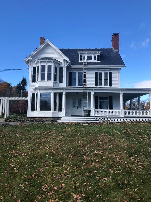 White two-story house with bay windows and a wrap-around porch, a ladder against the side, and a lawn under a blue sky.