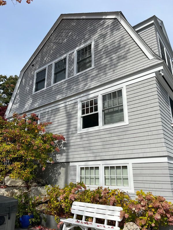 Gray shingled house with white-framed windows. A white bench sits near colorful flowering bushes.