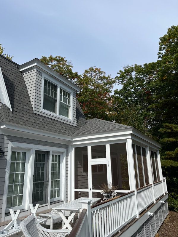 Gray shingle-clad house with screened porch and white trim, overlooking a deck and trees under a blue sky.