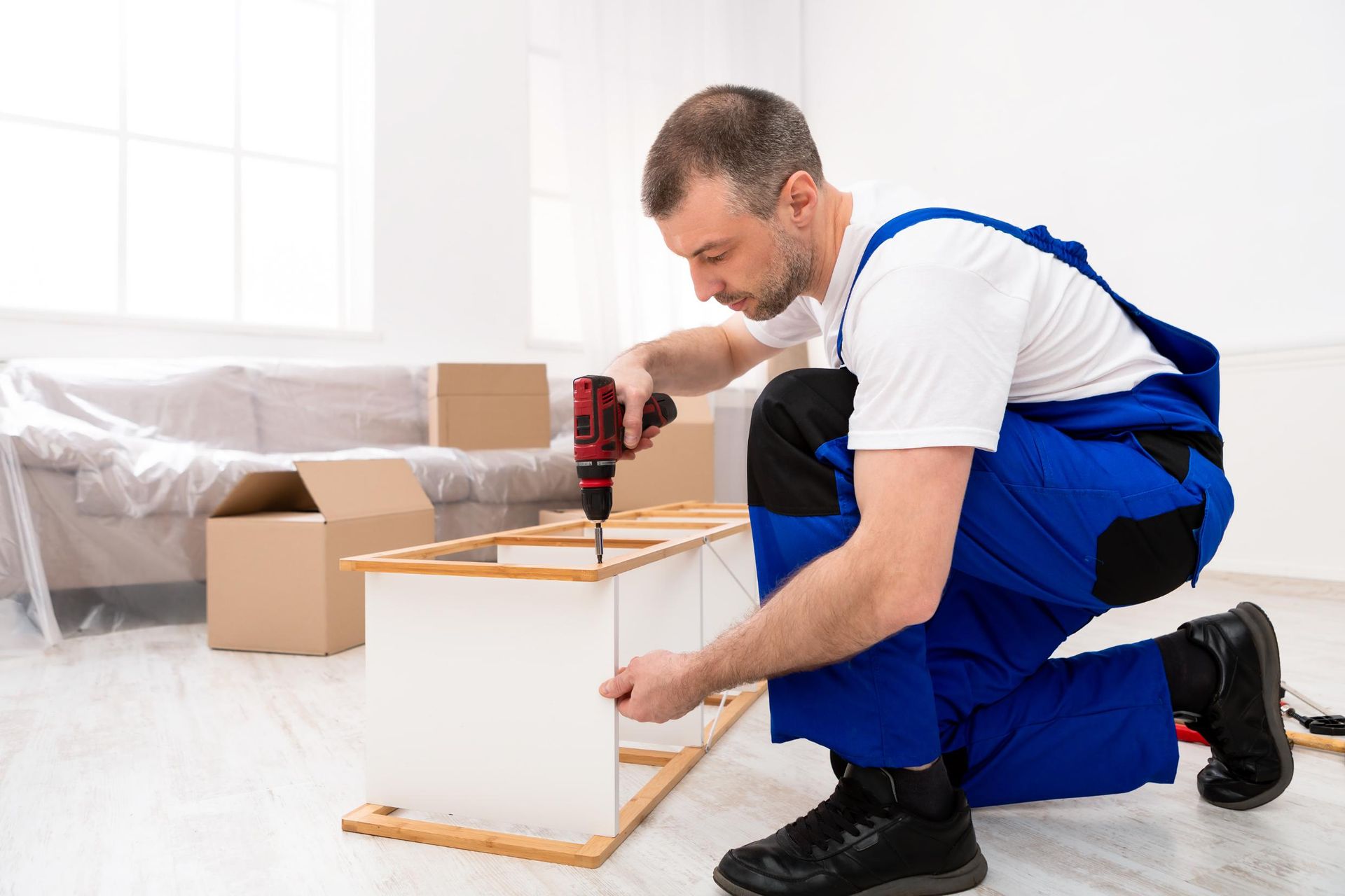 A man is kneeling down and using a drill to fix a piece of furniture.