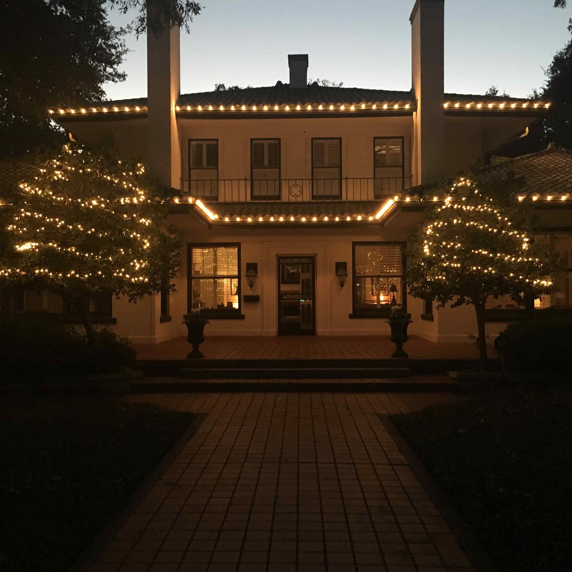 A house at dusk with string lights on the roof and trees, pathway leading to front door.