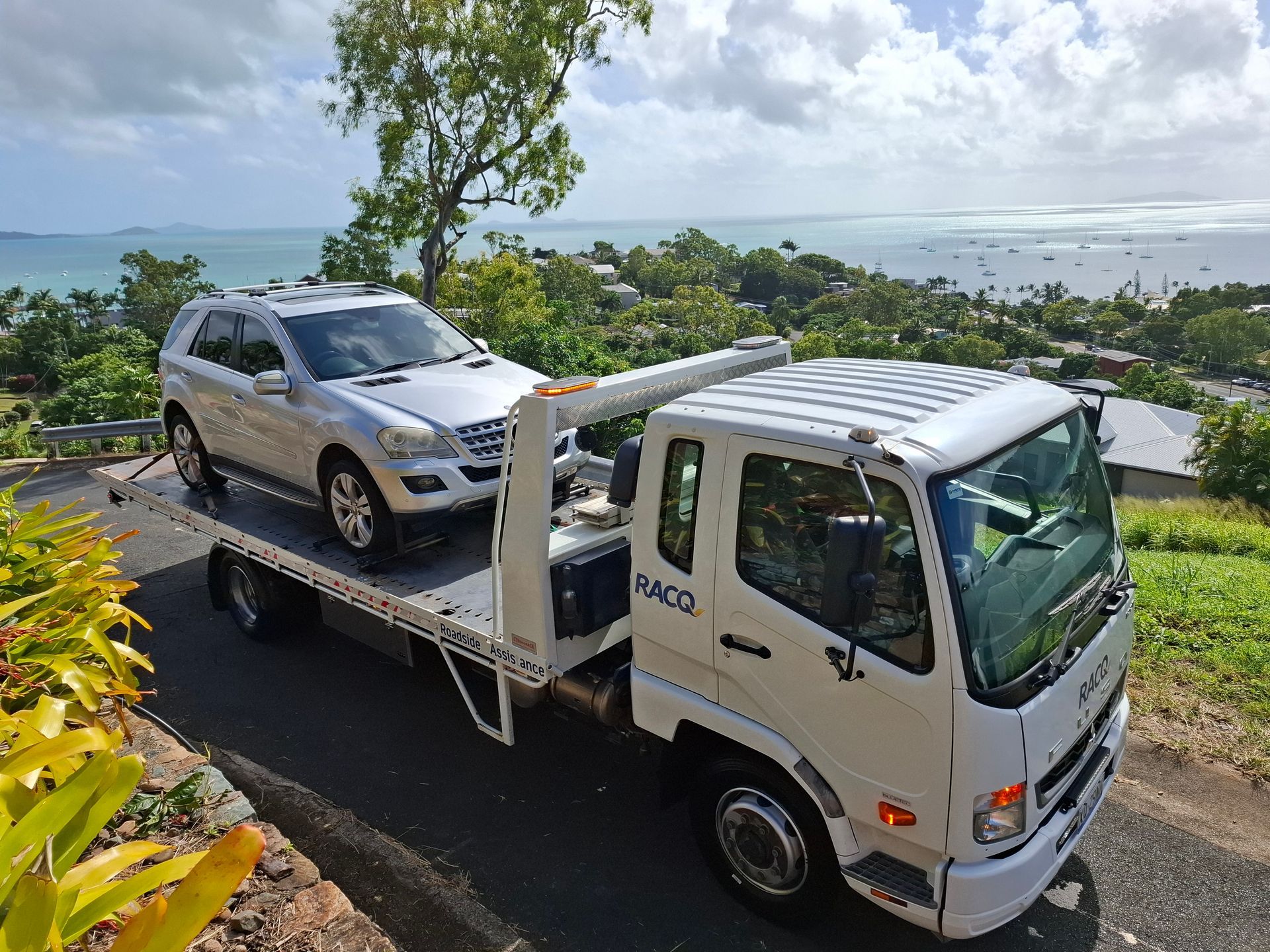 Tow truck carrying a silver SUV on a flatbed beside a coastal road with ocean view — Airlie Towing & Tilt Tray Hire In Cannonvale, QLD