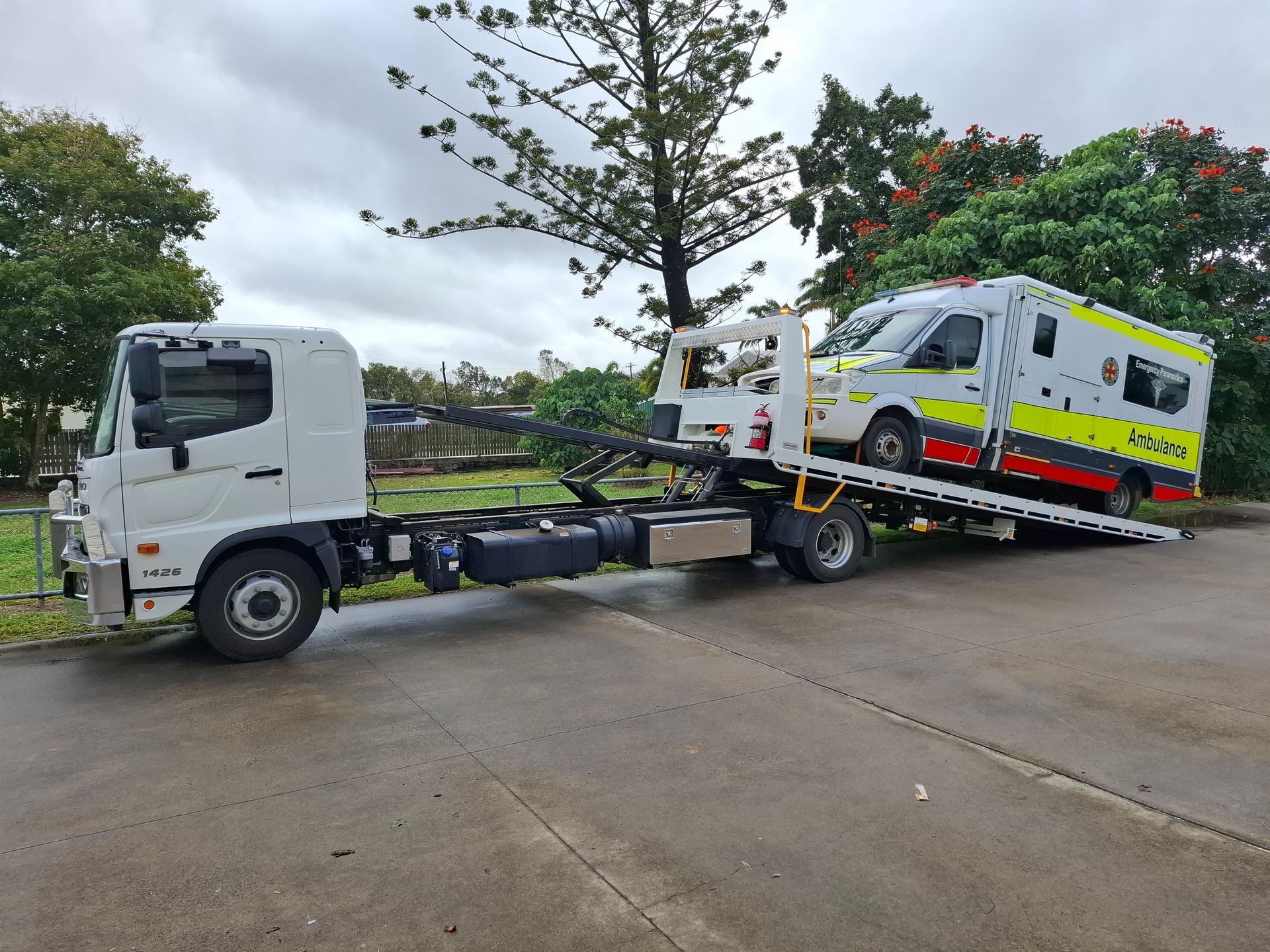 Tow truck carrying a white and yellow ambulance on a flatbed beside a wet road— Airlie Towing & Tilt Tray Hire In Airlie Beach, QLD