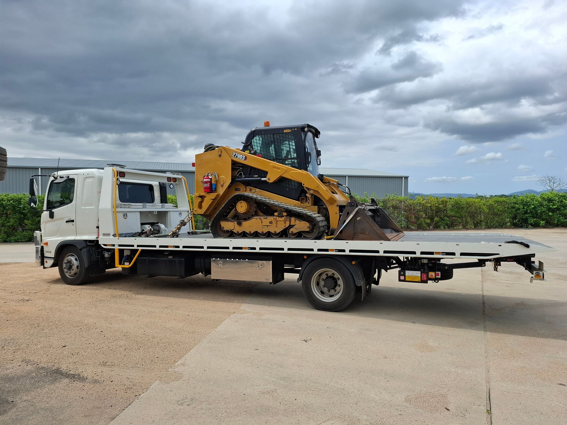 White flatbed tow truck carrying a yellow bulldozer on a rural roadside under cloudy skies — Airlie Towing & Tilt Tray Hire In Bowen, QLD