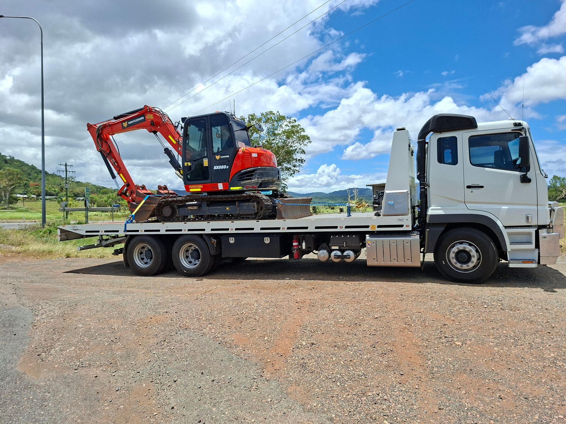 Flatbed truck carrying a red excavator on a gravel lot under a cloudy sky — Airlie Towing & Tilt Tray Hire In Cannonvale, QLD