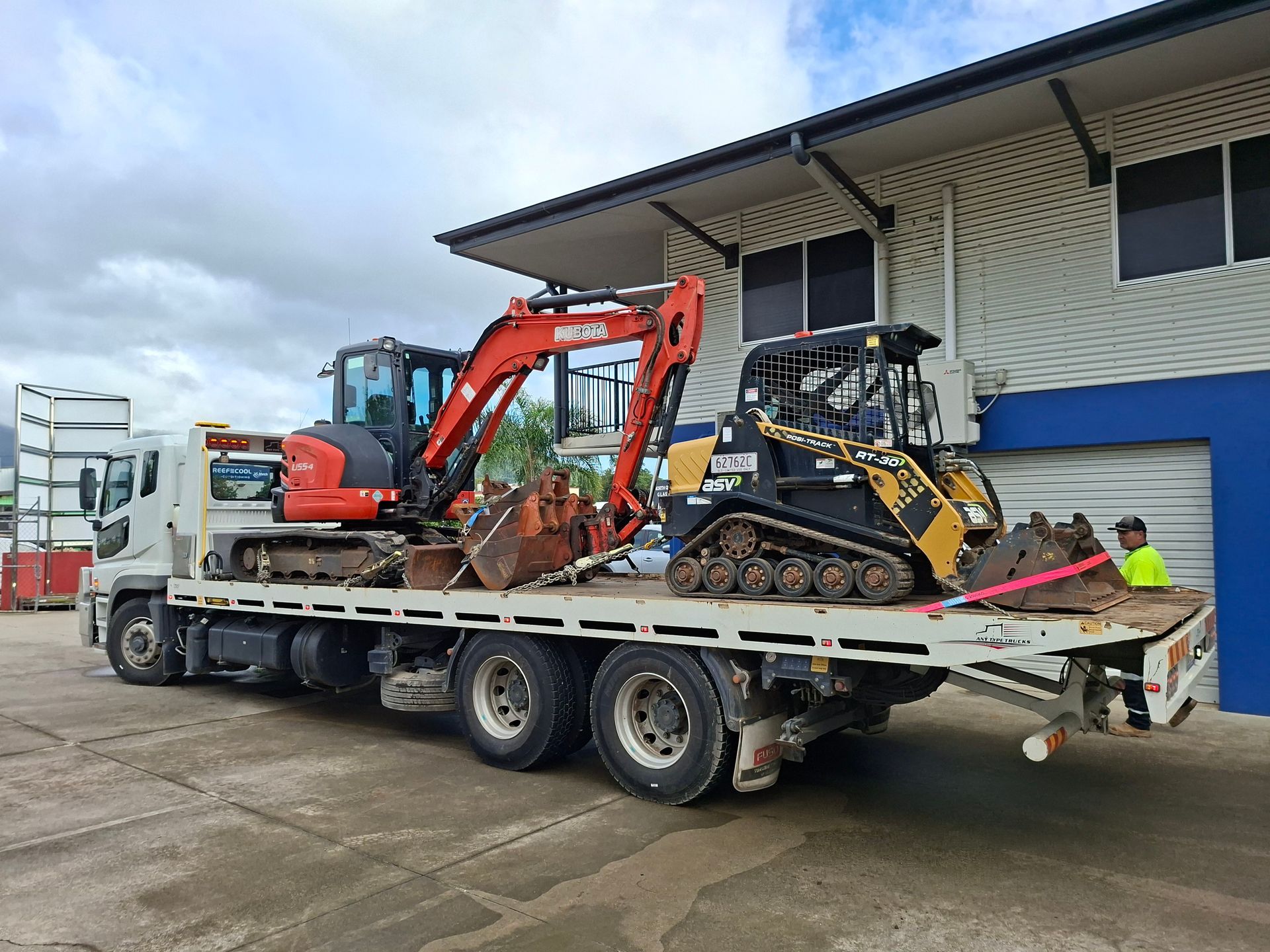 Flatbed truck carrying an orange excavator and yellow mini loader beside a blue warehouse — Airlie Towing & Tilt Tray Hire In Cannonvale, QLD