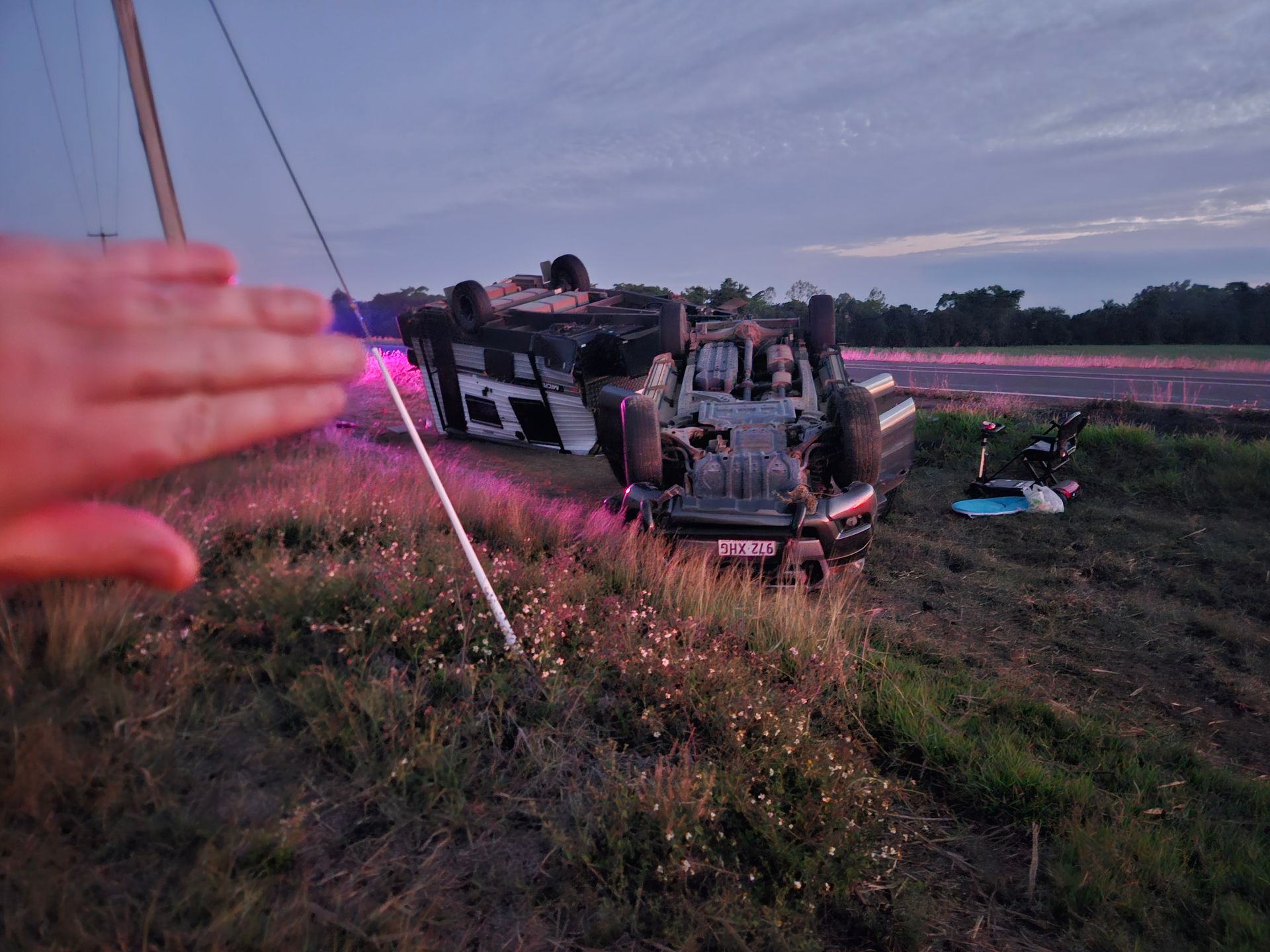 Overturned car in a roadside ditch at dusk, with a hand in the foreground and emergency lights glowing.— Airlie Towing & Tilt Tray Hire In Cannonvale, QLD