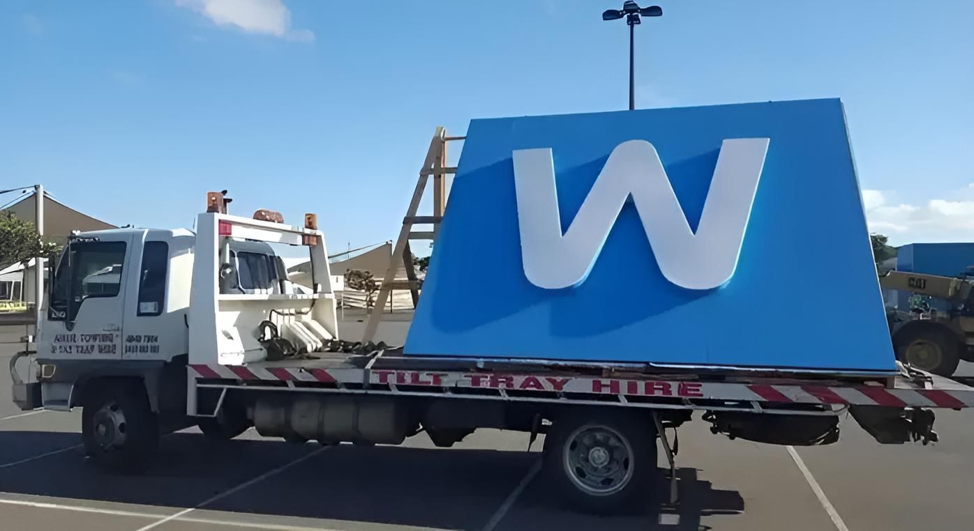 A Tow Truck is Carrying a Large Blue Sign With the Letter W on It — Airlie Towing & Tilt Tray Hire In Cannonvale, QLD