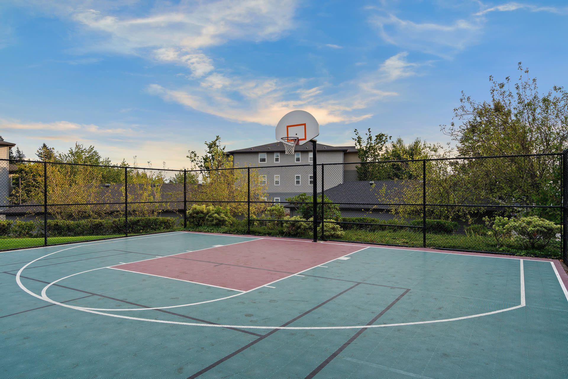 Outdoor basketball court with a hoop, chain-link fence, and trees.