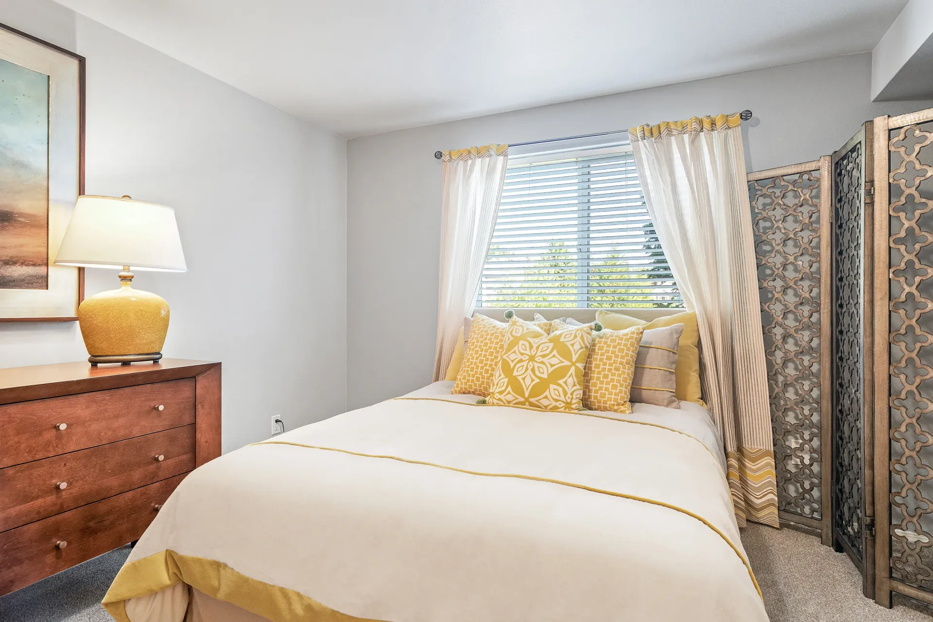 Bedroom in an apartment with a bed, dresser, curtains, window blinds, and a decorative folding screen.