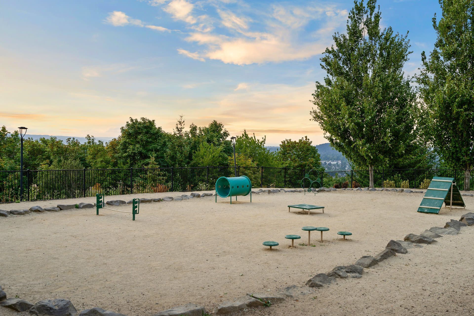 Outdoor community playground with green play equipment on sand, surrounded by trees and a fence.