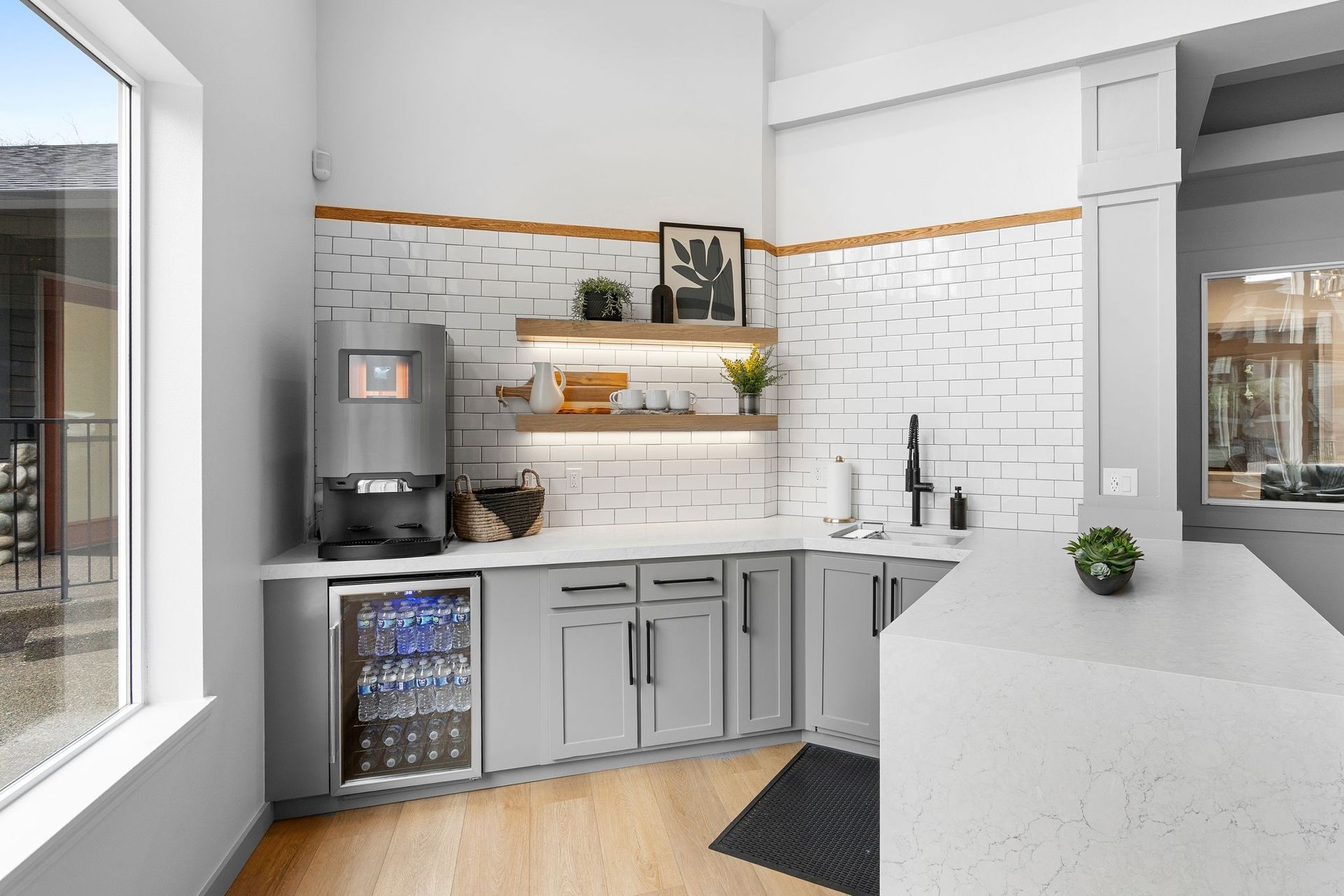 A modern kitchenette with gray cabinets, a beverage cooler, coffee machine, white brick backsplash, and a stone island.