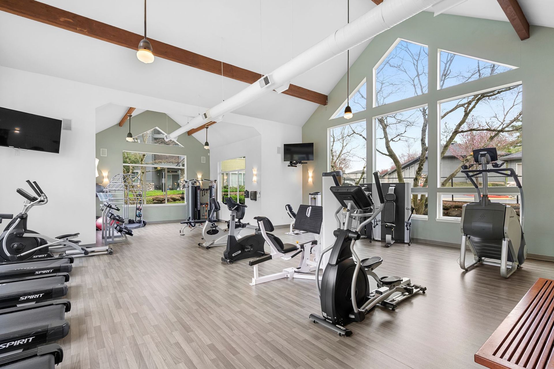 A well-lit, spacious apartment gym featuring various exercise machines, hardwood floors, and large windows.