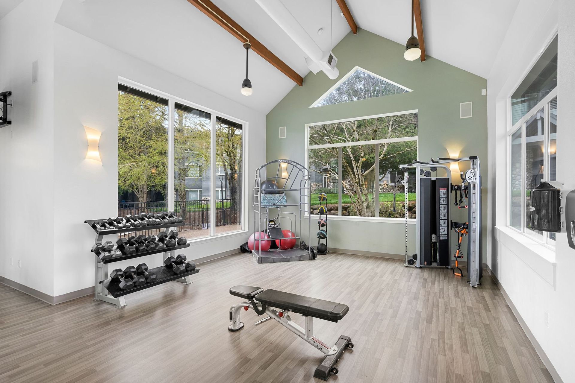 A bright, modern fitness center with wood-look flooring, a dumbbell rack, an adjustable workout bench, and gym equipment.