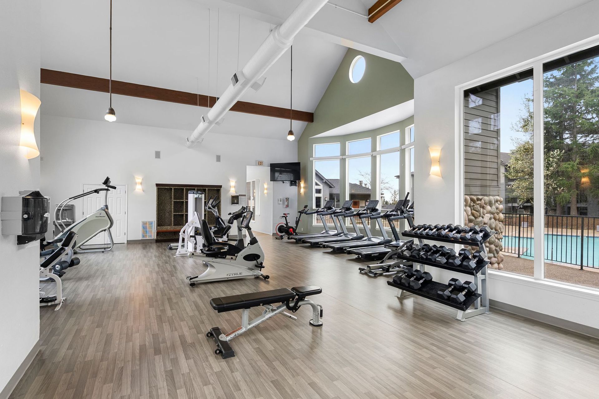 A well-lit, spacious fitness center with wooden flooring, exercise machines, treadmills, and a dumbbell weight rack.