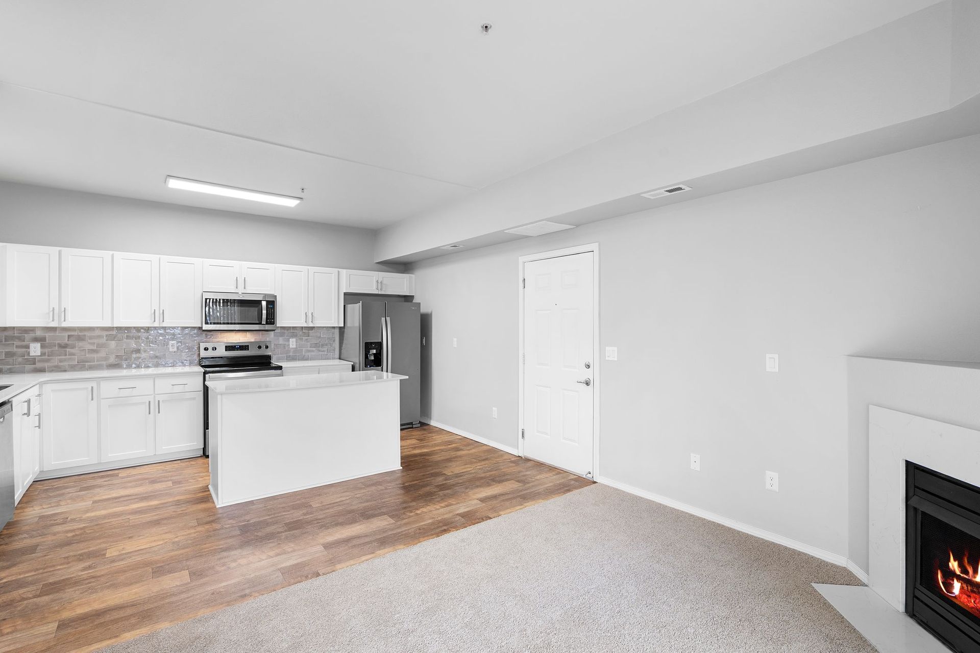 Open-concept kitchen and living area with white cabinets, light wood flooring, gray carpet, and a modern fireplace.