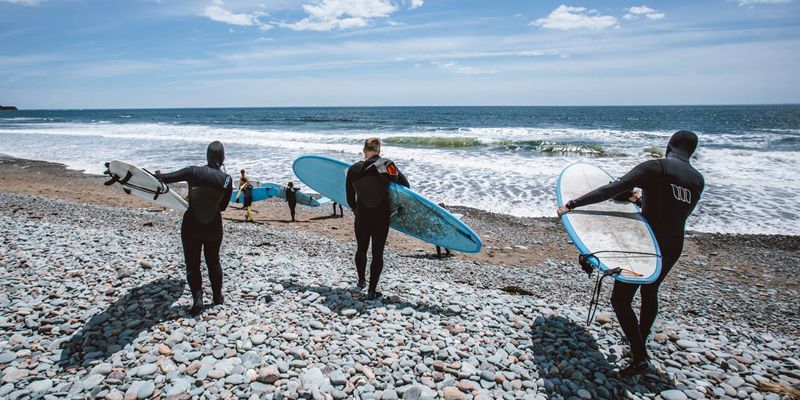 Three surfers are walking on a rocky beach carrying their surfboards.