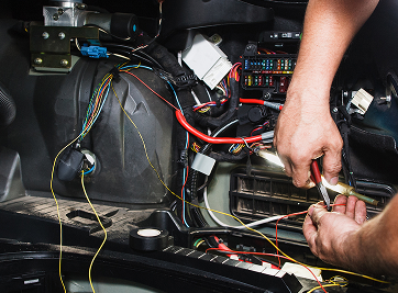 Person working on car wiring, using pliers on a red wire near a fuse box. Interior shot with visible wires. | Zee's Alignment & Autocare