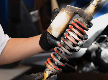 Person in black gloves holding a motorcycle shock absorber, showing a red spring and a silver body. | Zee's Alignment & Autocare
