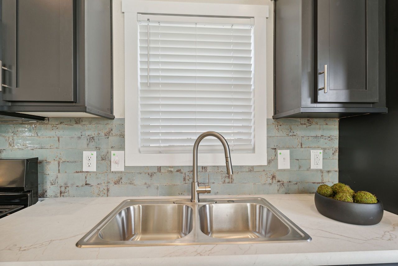 Kitchen sink with stainless steel double basins, white countertop, gray cabinets, blue tiled backsplash, window with blinds.
