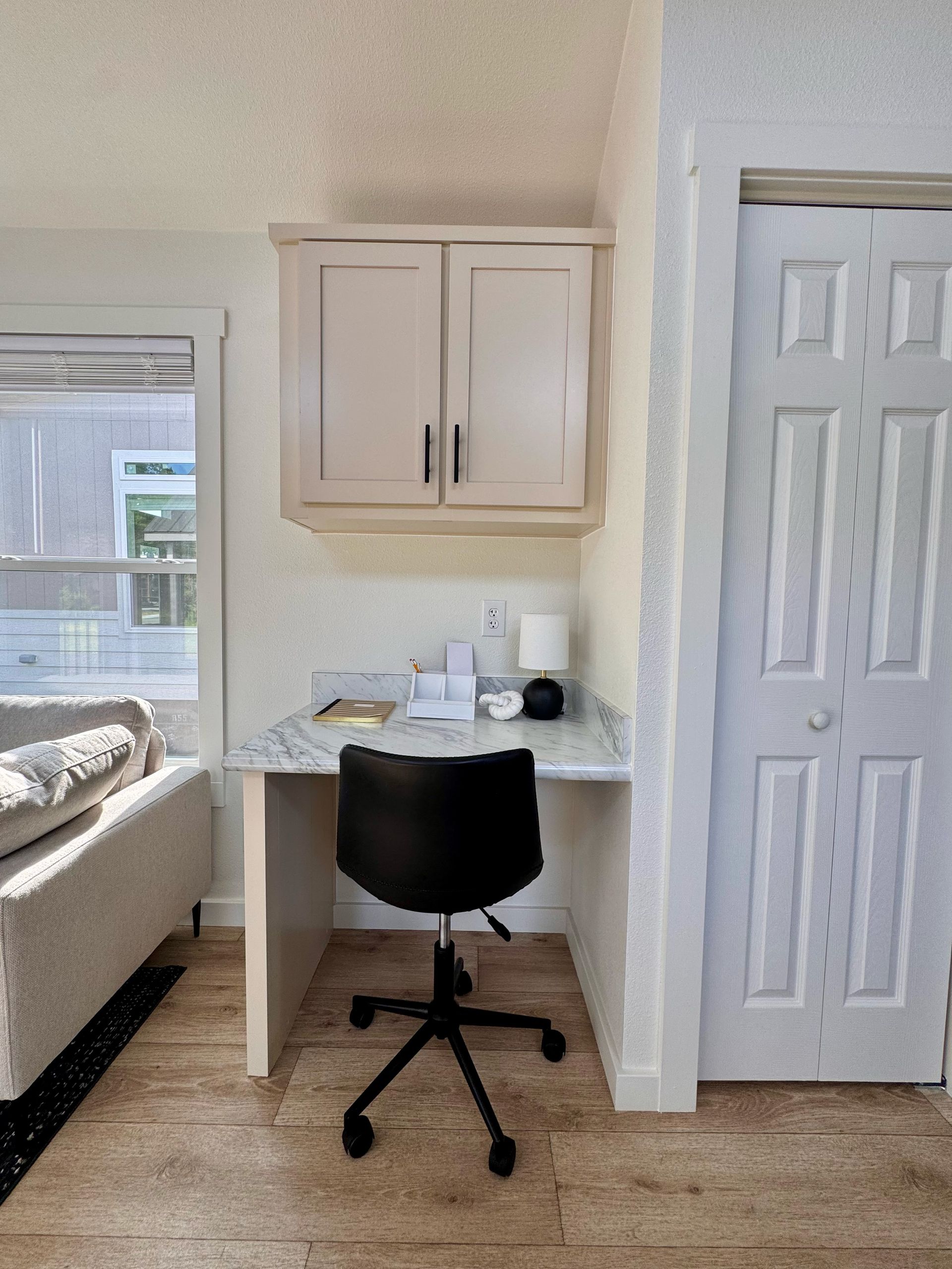 Small home office with a desk, chair, and overhead cabinets. The desk is between a window and a white door.